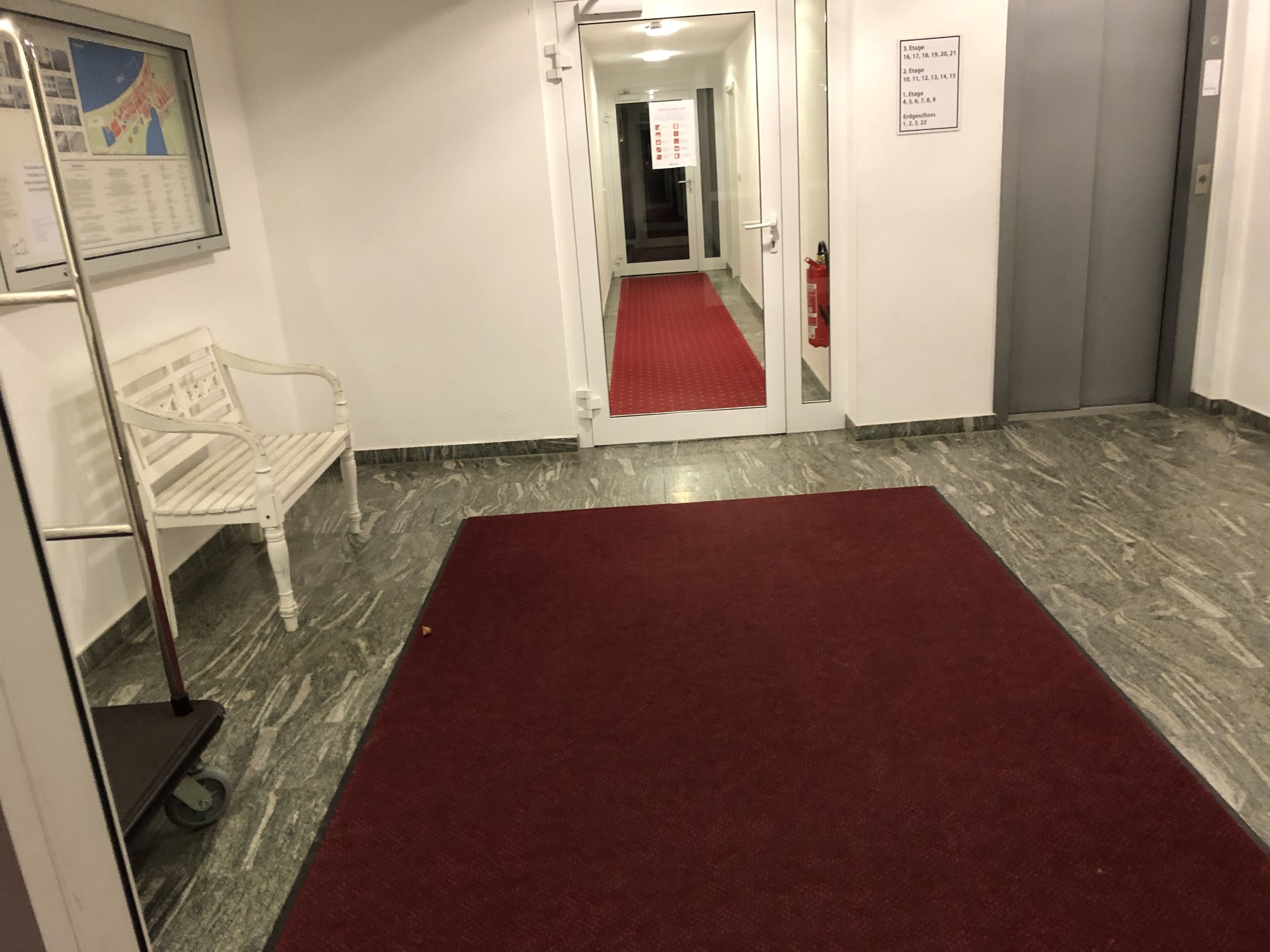 Un pasillo con alfombra roja, un banco blanco y una silla blanca, además de un cartel en la pared con los números de las habitaciones.