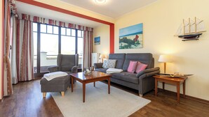 A living room with a sofa, armchair, coffee table, and lamps. There is a window with curtains, a painting on the wall, and a ship decoration.