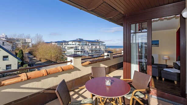 A balcony with a table and chairs, a view of buildings, and a glass door leading to a room with a sofa and a lamp.