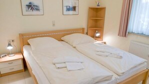 A double bed with white linens and towels, a bedside table with a lamp, and a shelf unit against the wall.