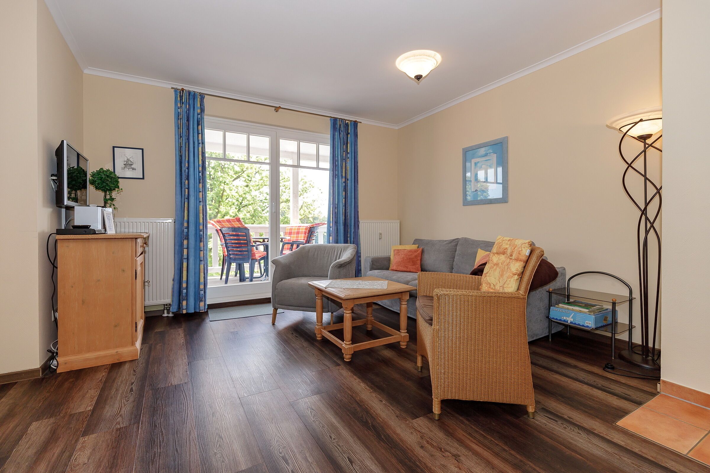 A living room with a wooden floor, a sofa, a chair, a table, a TV, a lamp, and a view of the outside.