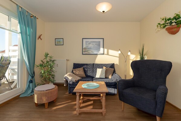 Ein Wohnzimmer mit einem dunkelblauen Sessel, einem gemusterten Sofa, einem runden Korbstuhl und einer Stehlampe.