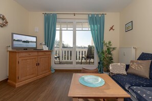 Un salon comprenant un meuble en bois, un téléviseur, un canapé, une table et une vue sur l’extérieur grâce à une grande fenêtre agrémentée de rideaux.