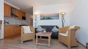 A modern kitchen with wooden cabinets, a microwave, and a coffee maker. There is a dining area with wicker chairs and a table, and a gray sofa with striped pillows. A painting of a beach scene is on the wall, and two floor lamps are on either side of the sofa.