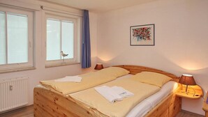 A wooden bed with two pillows and a blanket, a window with a curtain, a wall-mounted lamp, and a framed picture.
