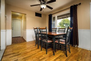 A dining room with a wooden table and chairs, a ceiling fan, and a window with curtains.