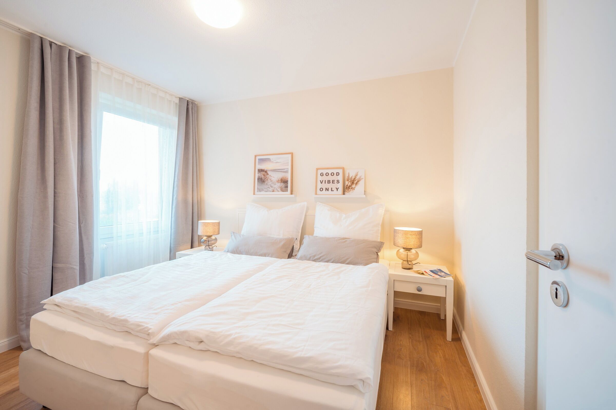 A hotel room with a double bed, two bedside tables with lamps, a window with curtains, and two framed pictures on the wall.