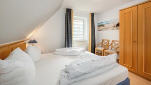 Un dormitorio con cama, mesita de noche, lámpara, ventana con cortinas, una silla y armario.