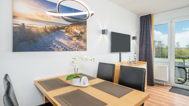 Una habitación moderna con una mesa de comedor, sillas, un televisor montado, un ventanal con vistas y una lámpara colgante.
