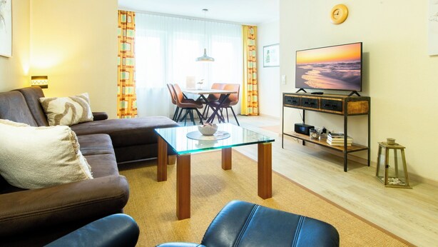 Una sala de estar con un sofá, una mesa de centro de vidrio, un televisor sobre un mueble de madera y un comedor con sillas y una mesa.