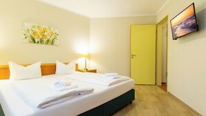 Una habitación de hotel con una cama grande, dos almohadas, una mesita de noche con una lámpara, un cuadro en la pared y un televisor montado.