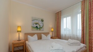 A neatly made bed with white linens and towels, two bedside lamps, a painting of a flower on the wall, and a window with curtains.