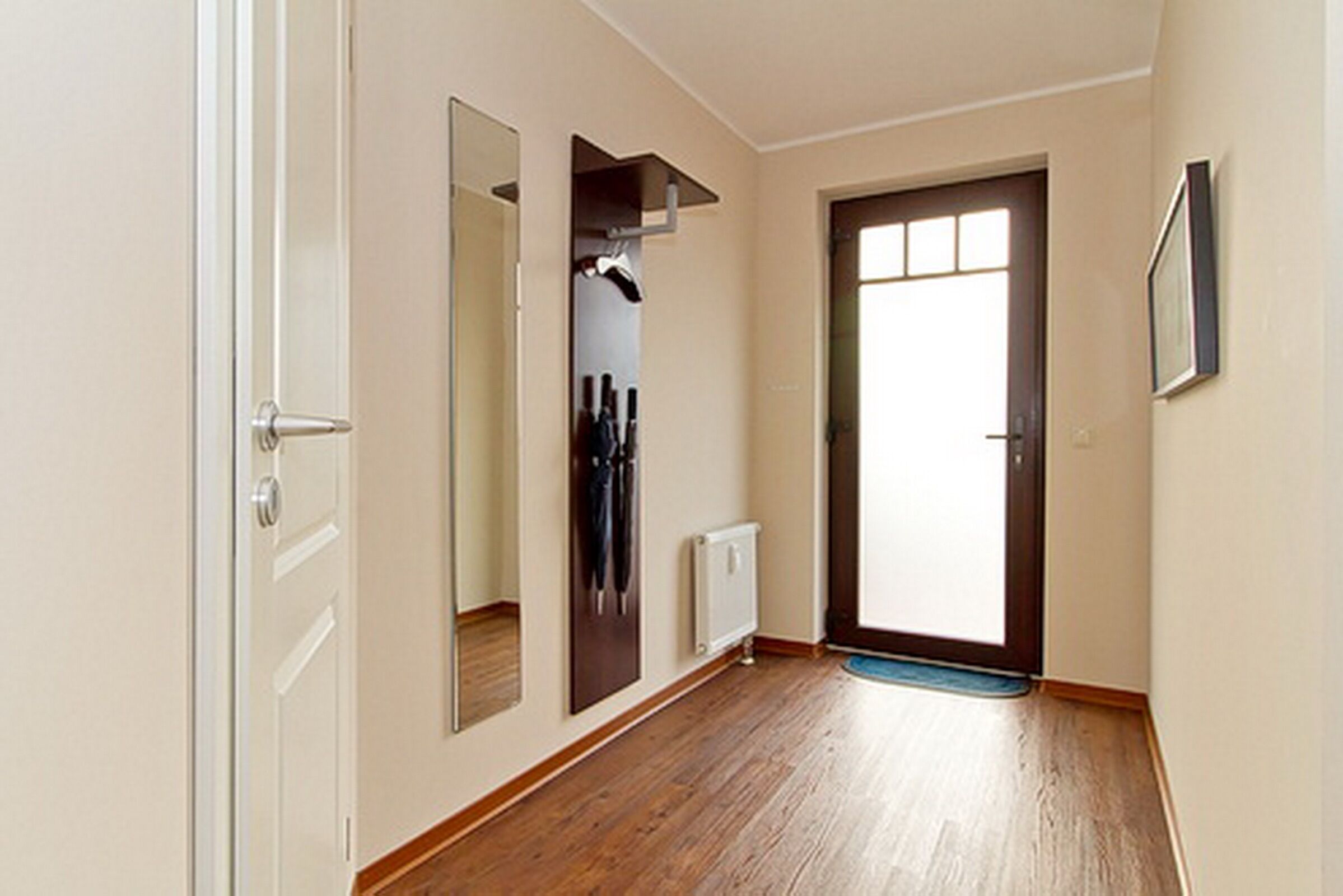Un couloir avec un plancher en bois, une porte blanche, un miroir, un radiateur et un tableau encadré accroché au mur.