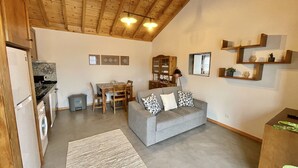 Una sala de estar acogedora con un sofá gris, vigas de madera en el techo, un comedor con mesa y sillas, y una cocina equipada con refrigerador y lavadora.
