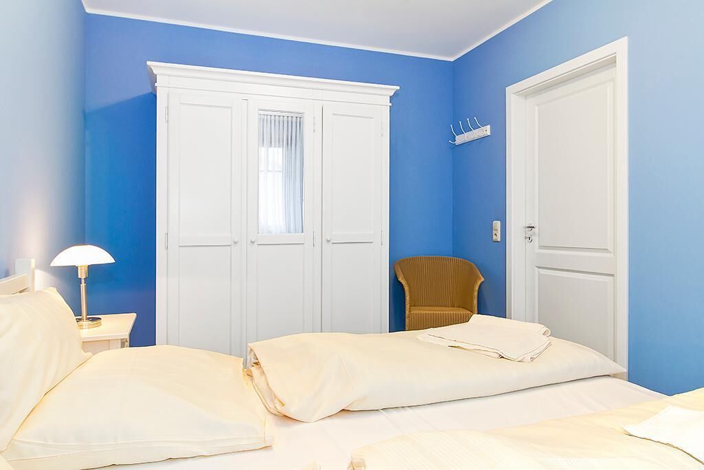 A bedroom with two beds, a white wardrobe, a chair, and a nightstand with a lamp.