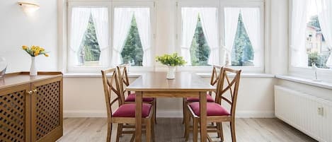 En matplats med ett träbord och stolar, stora fönster med gardiner och utsikt över träd utanför.