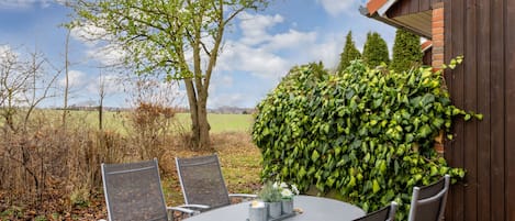 An outdoor patio with a round table and four chairs, surrounded by a hedge and a wooden fence.