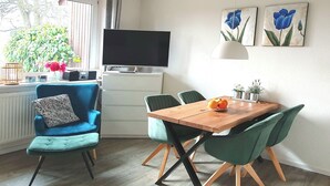 A modern living room with a TV, a wooden dining table, green chairs, a blue armchair, and a window with plants.