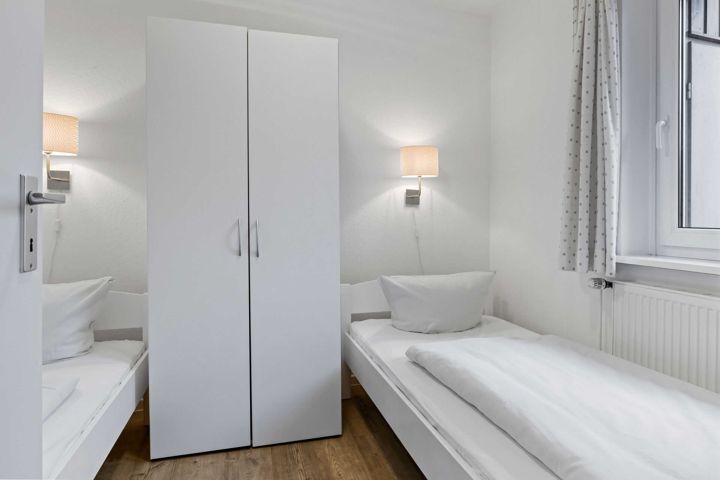 A bedroom with a double bed, a wardrobe, a bedside table, a lamp, and a window with curtains.