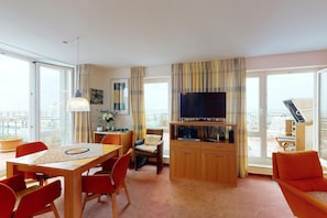 Uma sala de estar moderna com uma mesa de jantar de madeira, cadeiras vermelhas, uma TV sobre um suporte de madeira e janelas amplas com vista para o mar.