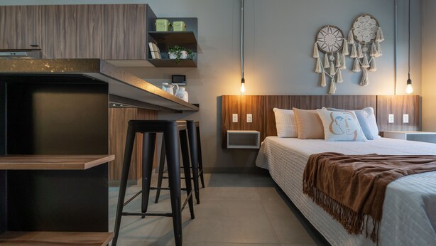 Una habitación de hotel moderna con kitchenette, una cama con manta marrón, dos atrapasueños colgados en la pared y una lámpara colgante.