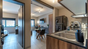 Una cocina moderna con refrigerador empotrado, cafetera y pileta. Se ve un comedor con mesa y sillas a través de un umbral abierto.