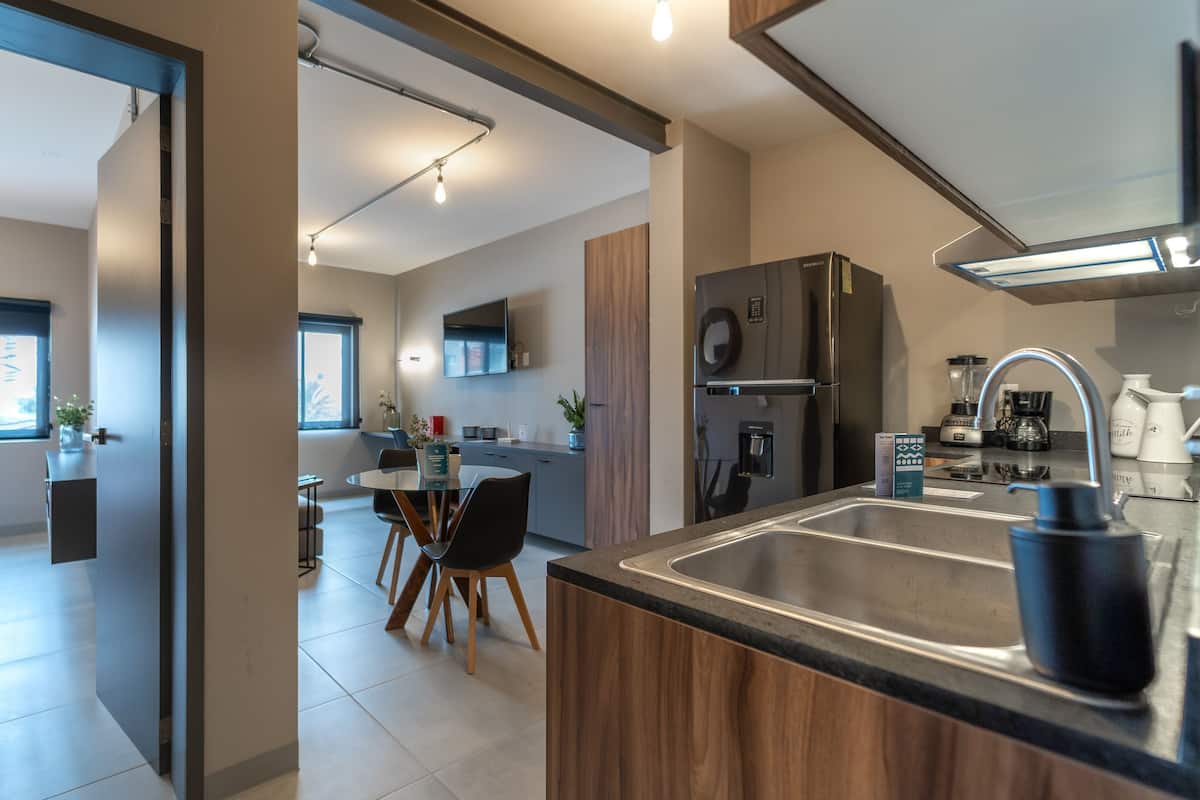 Una cocina moderna con refrigerador empotrado, cafetera y pileta. Se ve un comedor con mesa y sillas a través de un umbral abierto.