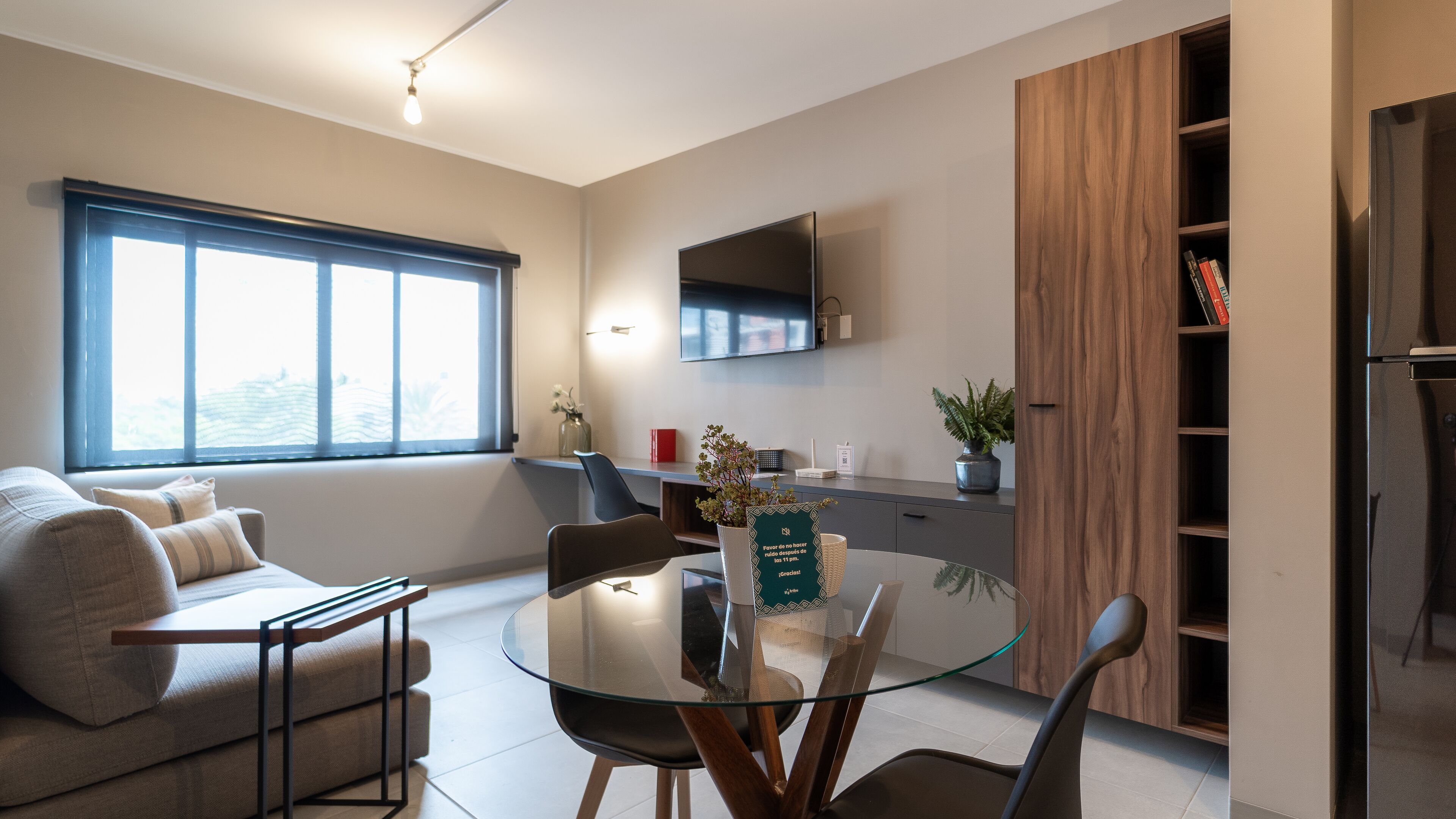 Een moderne woonkamer met een glazen eettafel, bank en een flatscreen televisie aan de muur.