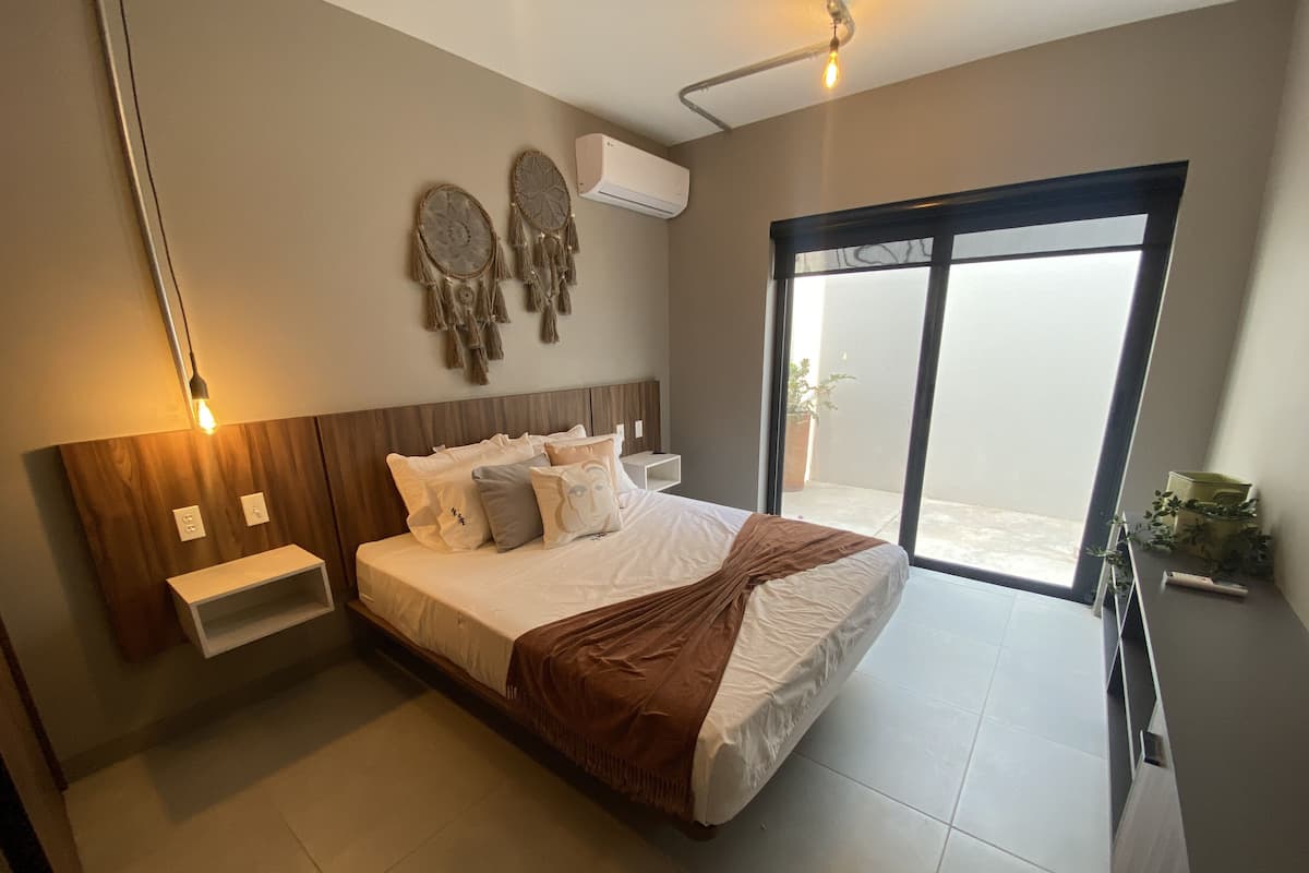 Un dormitorio moderno con una cama grande, mesitas de noche, aire acondicionado instalado en la pared y un ventanal corredero al balcón.