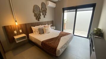 Un dormitorio moderno con una cama grande, mesitas de noche, aire acondicionado instalado en la pared y un ventanal corredero al balcón.