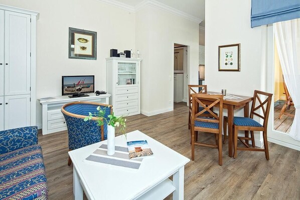A living room with a television, a dining table with chairs, a blue patterned armchair, and a white cabinet.
