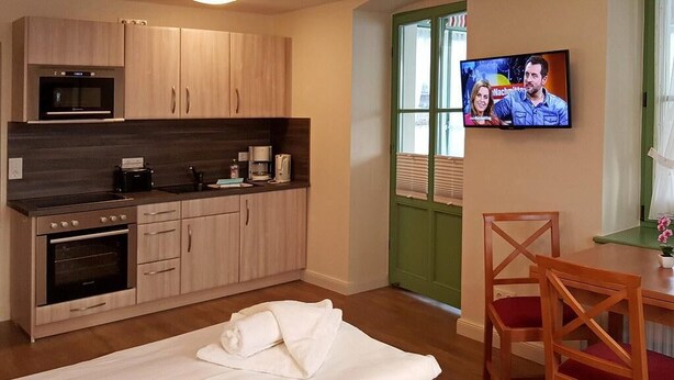 A compact room with a kitchenette, a bed with white linens, a television mounted on the wall, and a wooden chair with red cushions.