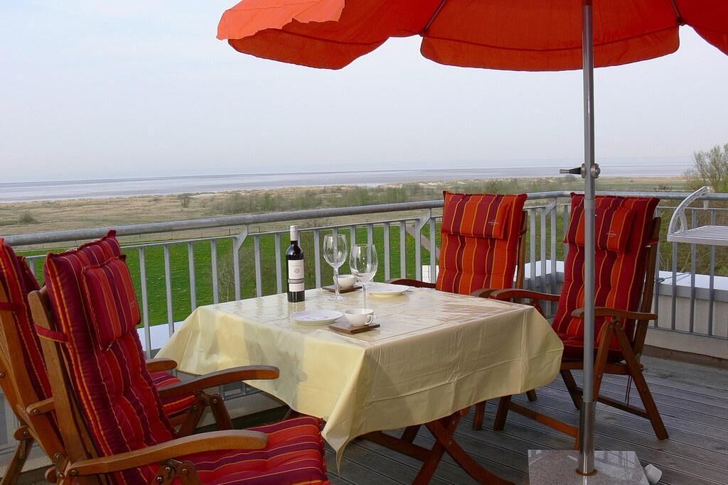 Una mesa dispuesta con una botella, vasos y una taza en un balcón con vista a la playa.