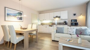 Una sala de estar moderna y luminosa con comedor, una cocina con armarios blancos y un sofá decorado con cojines.