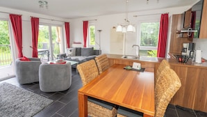 A modern kitchen with a wooden table and chairs, a dining area with a sofa and ottoman, and a balcony with red curtains. - Apartment mit Sauna, Whirlbadewanne und Balkon an der Graft mit Blick zum Deich! (Nordseeheilbad Schillig)