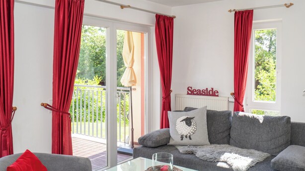 A room with a grey sofa, a glass coffee table, red curtains, and a 'Seaside' sign. - Apartment mit Sauna, Whirlbadewanne und Balkon an der Graft mit Blick zum Deich! (Nordseeheilbad Schillig)