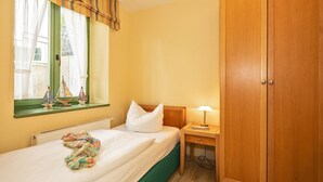A hotel room with a bed, a nightstand with a lamp, a wooden wardrobe, a window with curtains, and a small sailboat decoration on the windowsill.