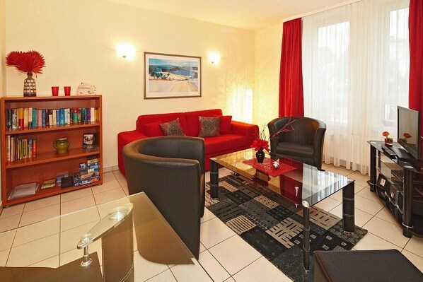 A living room with a red sofa, a black armchair, a glass coffee table, a bookshelf, and a painting on the wall.