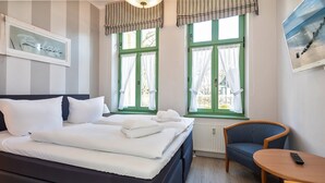 A neatly made bed with white linens, a blue upholstered chair, a small wooden table, and a framed picture on the wall. - 4-zimmer Ferienwohnung mit Wintergarten und Terrasse in Toplage an der Promenade (Heringsdorf)