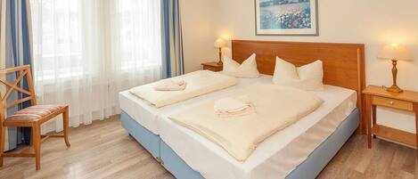 Una cama doble con cabecera de madera, dos mesitas de noche con lámparas, una silla y un gran ventanal con cortinas traslúcidas.