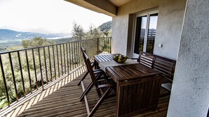 A balcony with a wooden table and chairs, a bowl of fruit, and a view of a valley and mountains. - New accommodation, summer kitchen, terrace, garden (Olmeto)