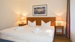 Una cama bien hecha con sábanas y almohadas blancas, dos lámparas de noche, un cuadro en la pared y un cabecero de madera.
