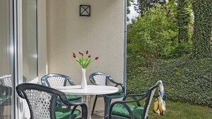 Un patio exterior con una mesa redonda y cuatro sillas, un florero con flores rojas y un seto al fondo.