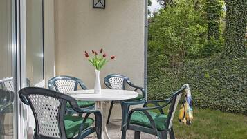Eine Terrasse im Freien mit einem runden Tisch und vier Stühlen, einer Vase mit roten Blumen und einem Hecken im Hintergrund.