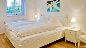 A bedroom with a white bed, two bedside tables with lamps, a window with a view of trees, and a painting on the wall.