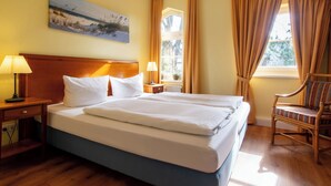 Una habitación de hotel con una cama grande, dos mesitas de noche con lámparas, una silla de madera y un cuadro en la pared.