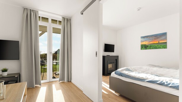 A modern bedroom with a bed, a TV, a fireplace, and a sliding door to a balcony. - Hübsche, Strandnahe Ferienwohnung mit Eigener Sauna und Terrasse mit Strandkorb (Nordseebad Wremen)