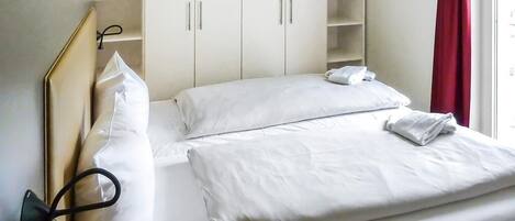 A hotel room with a bed, white bedding, a headboard, a shelf unit, and a red curtain.