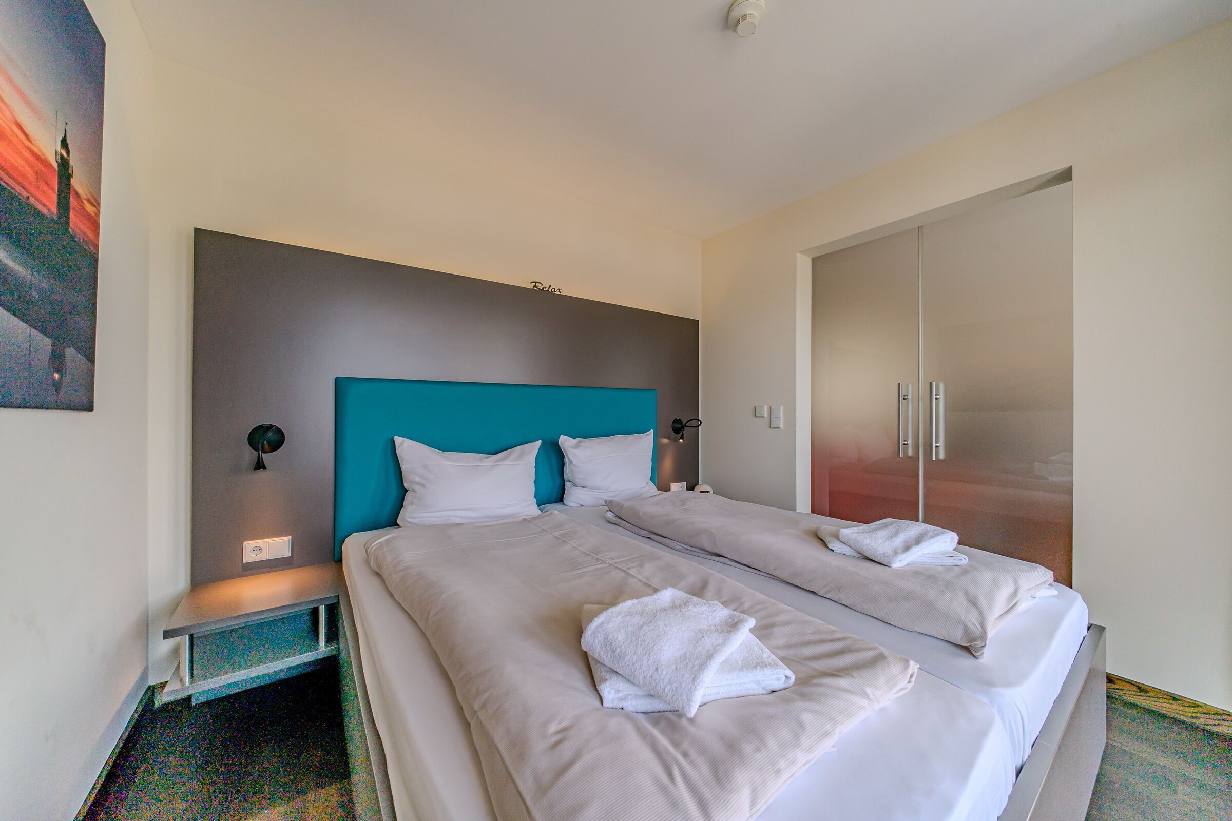 A hotel room with two beds, a teal headboard, white bedding, and a large wardrobe.