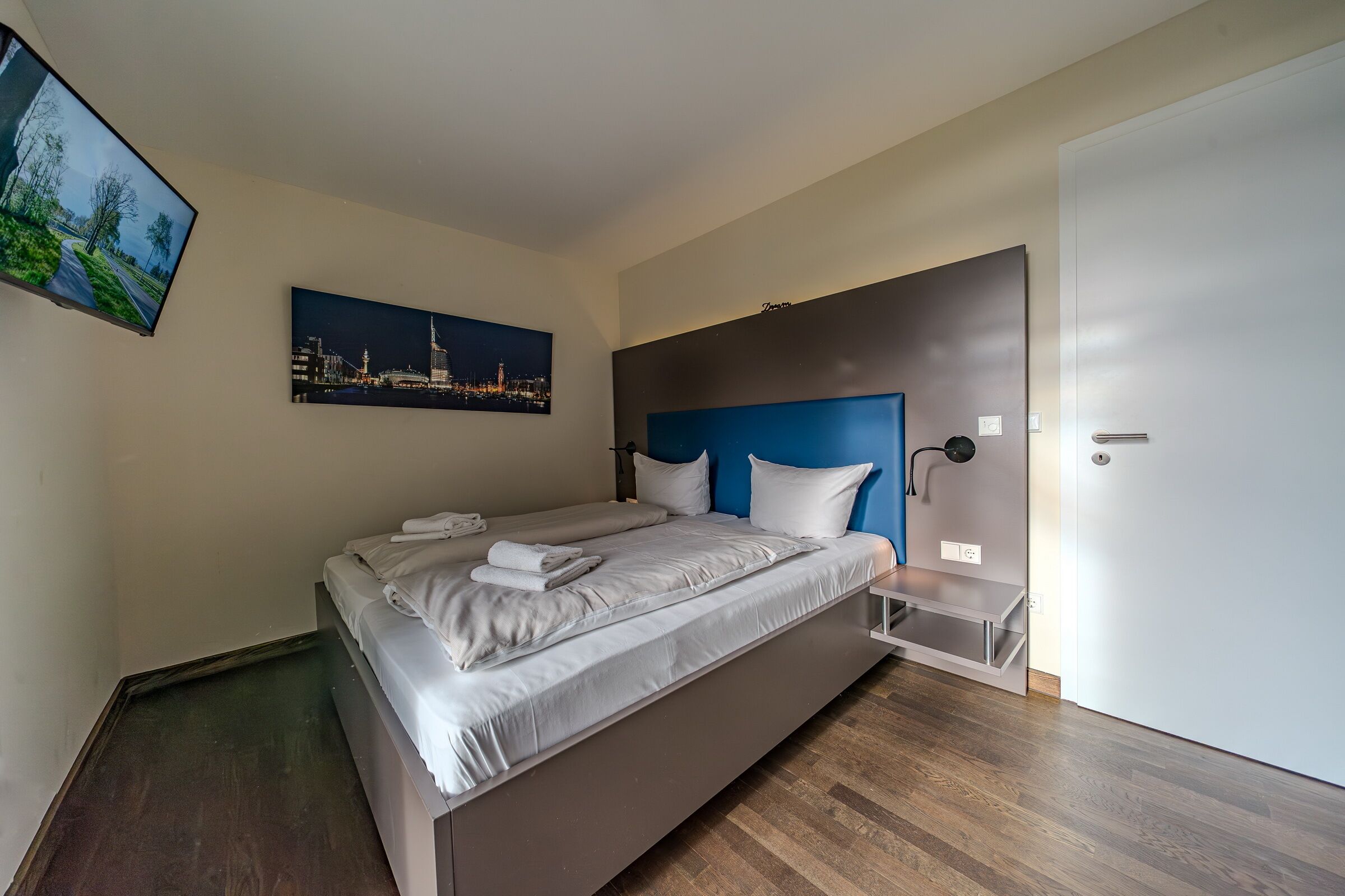 A modern hotel room with a large bed, a cityscape painting, a blue accent wall, and a wooden floor.